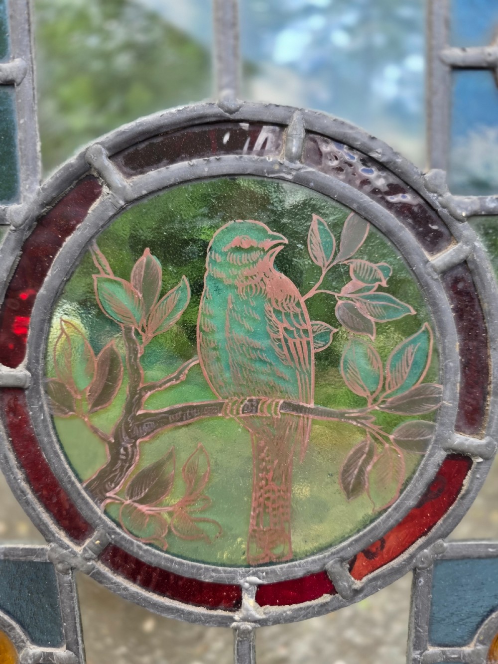 A square leaded stained glass panel featuring a central circular painted yellow bird on a branch, four amber bullseye roundels, and a light wood frame. View V