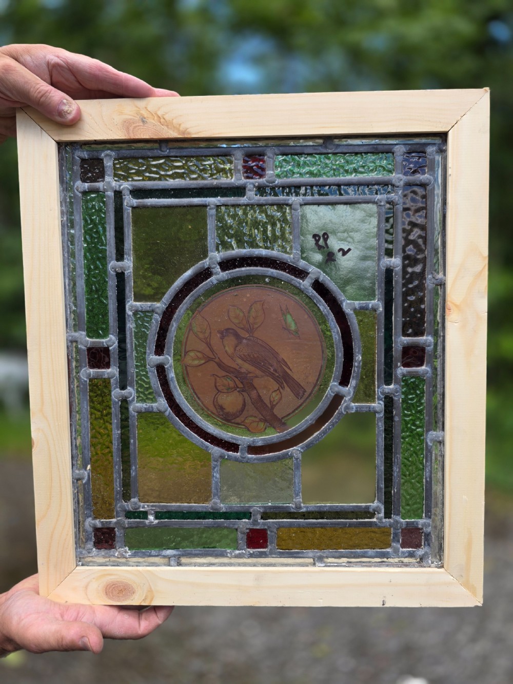 A square Edwardian leaded glass panel featuring a central hand-painted roundel of a bird on a branch surrounded by geometric green and amber glass within a timber frame.