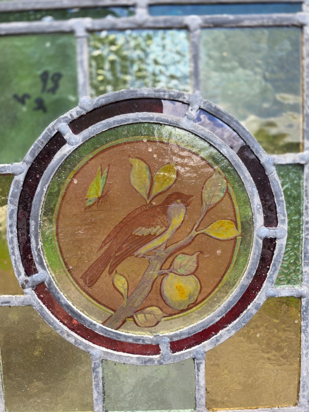 A square Edwardian leaded glass panel featuring a central hand-painted roundel of a bird on a branch surrounded by geometric green and amber glass within a timber frame. View IV