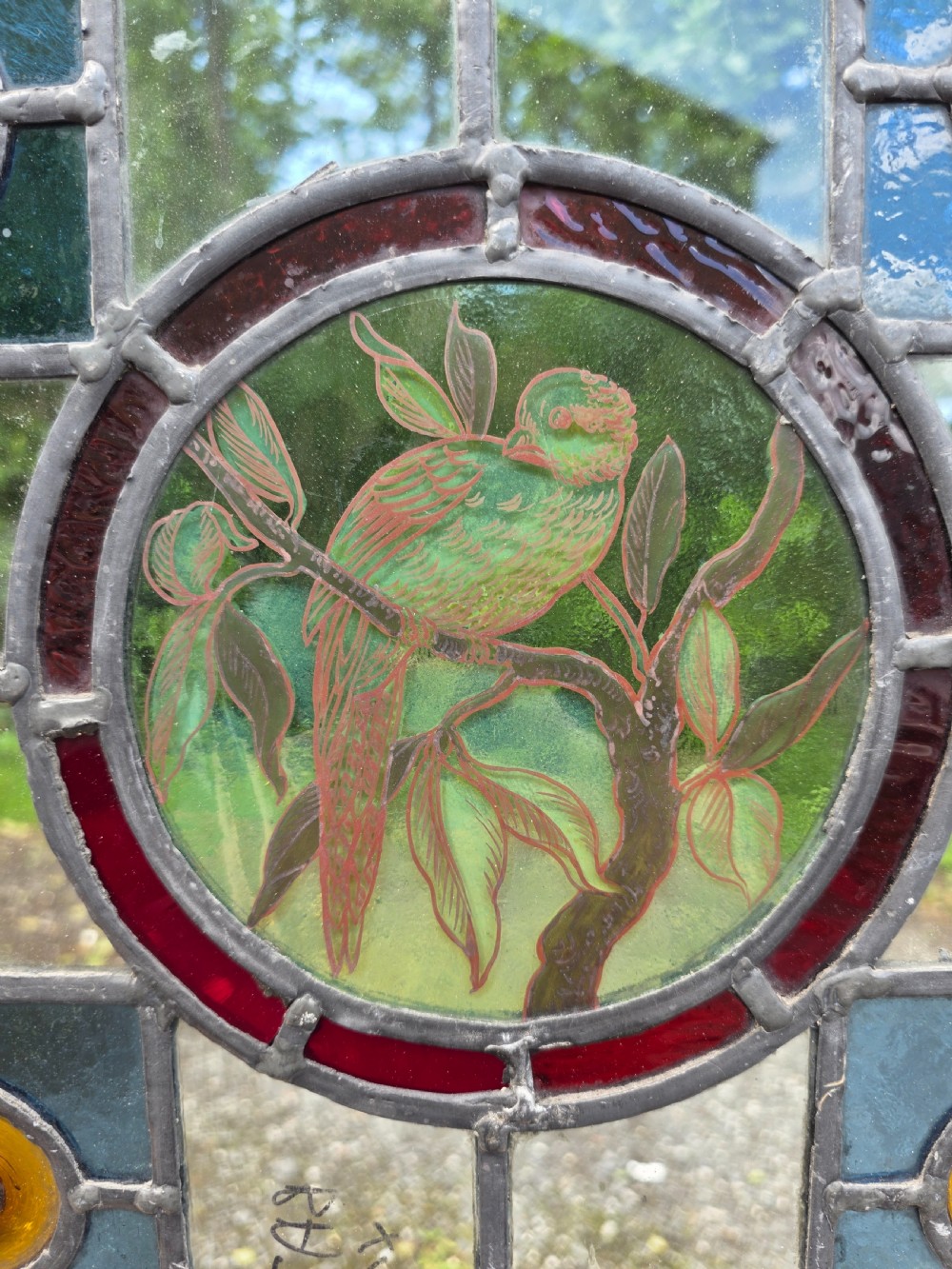 A square Edwardian leaded stained glass window featuring a central circular roundel painted with a green bird on a branch, surrounded by four amber bullseyes. View II