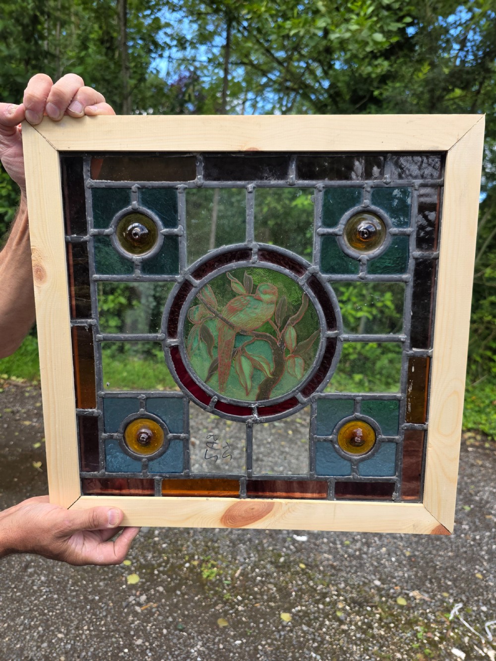 A square Edwardian leaded stained glass window featuring a central circular roundel painted with a green bird on a branch, surrounded by four amber bullseyes. View III