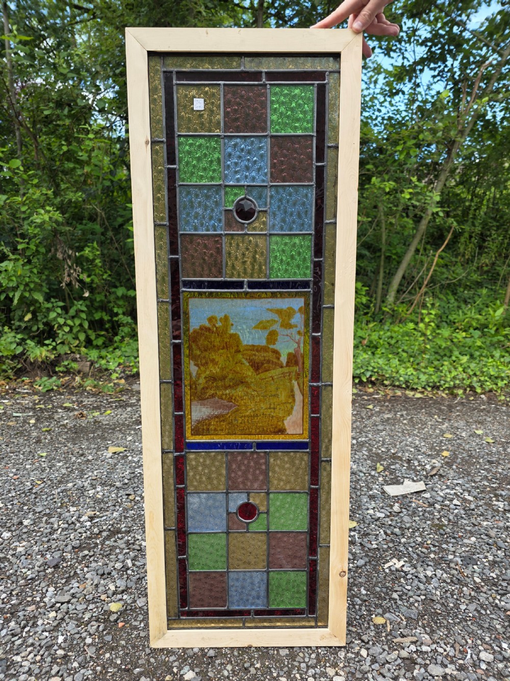 A narrow rectangular leaded glass window panel with geometric coloured panes, glass jewels, and a central hand-painted countryside scene.