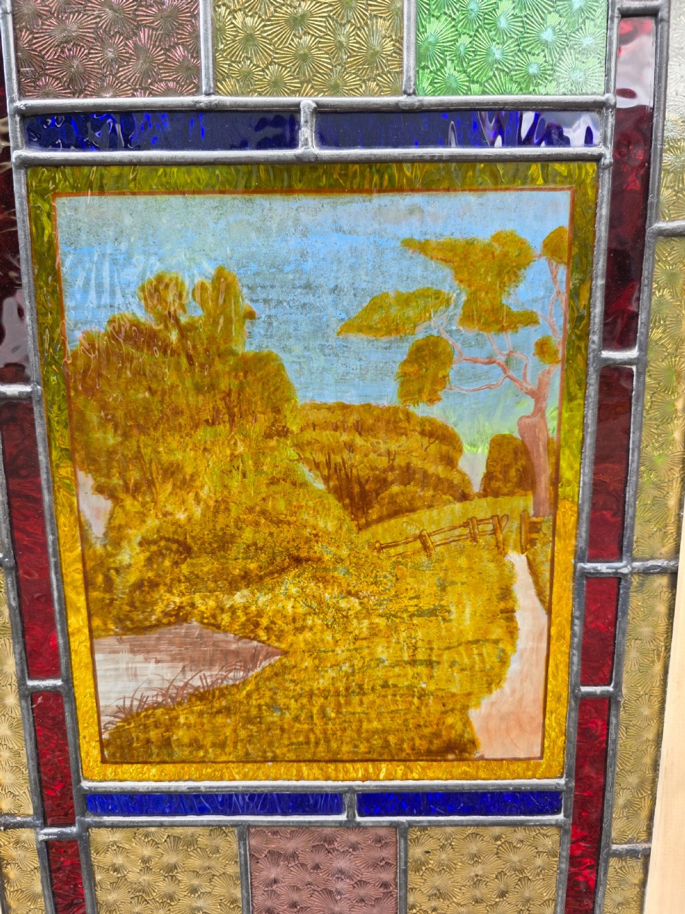 A narrow rectangular leaded glass window panel with geometric coloured panes, glass jewels, and a central hand-painted countryside scene. View II