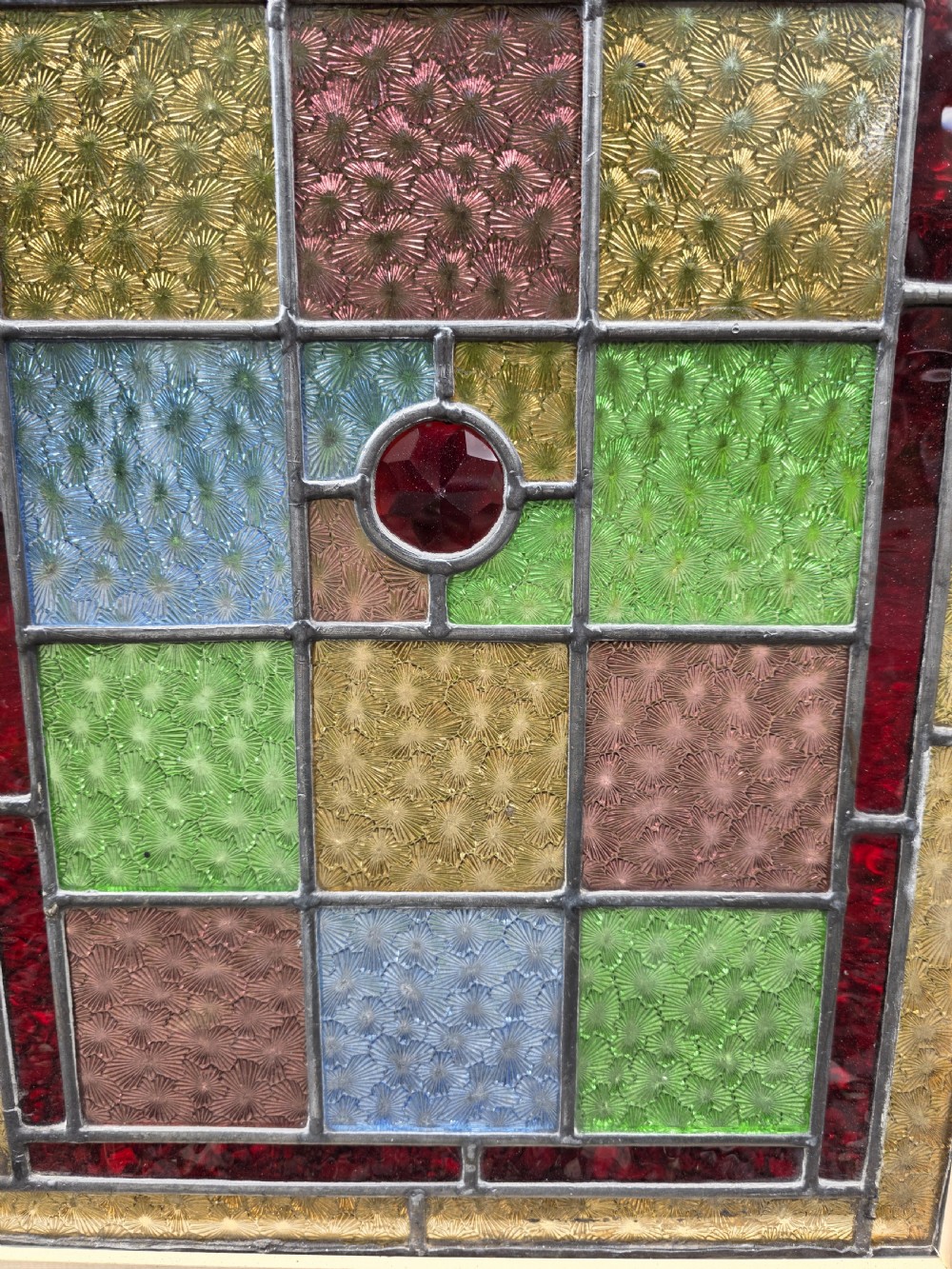 A narrow rectangular leaded glass window panel with geometric coloured panes, glass jewels, and a central hand-painted countryside scene. View III