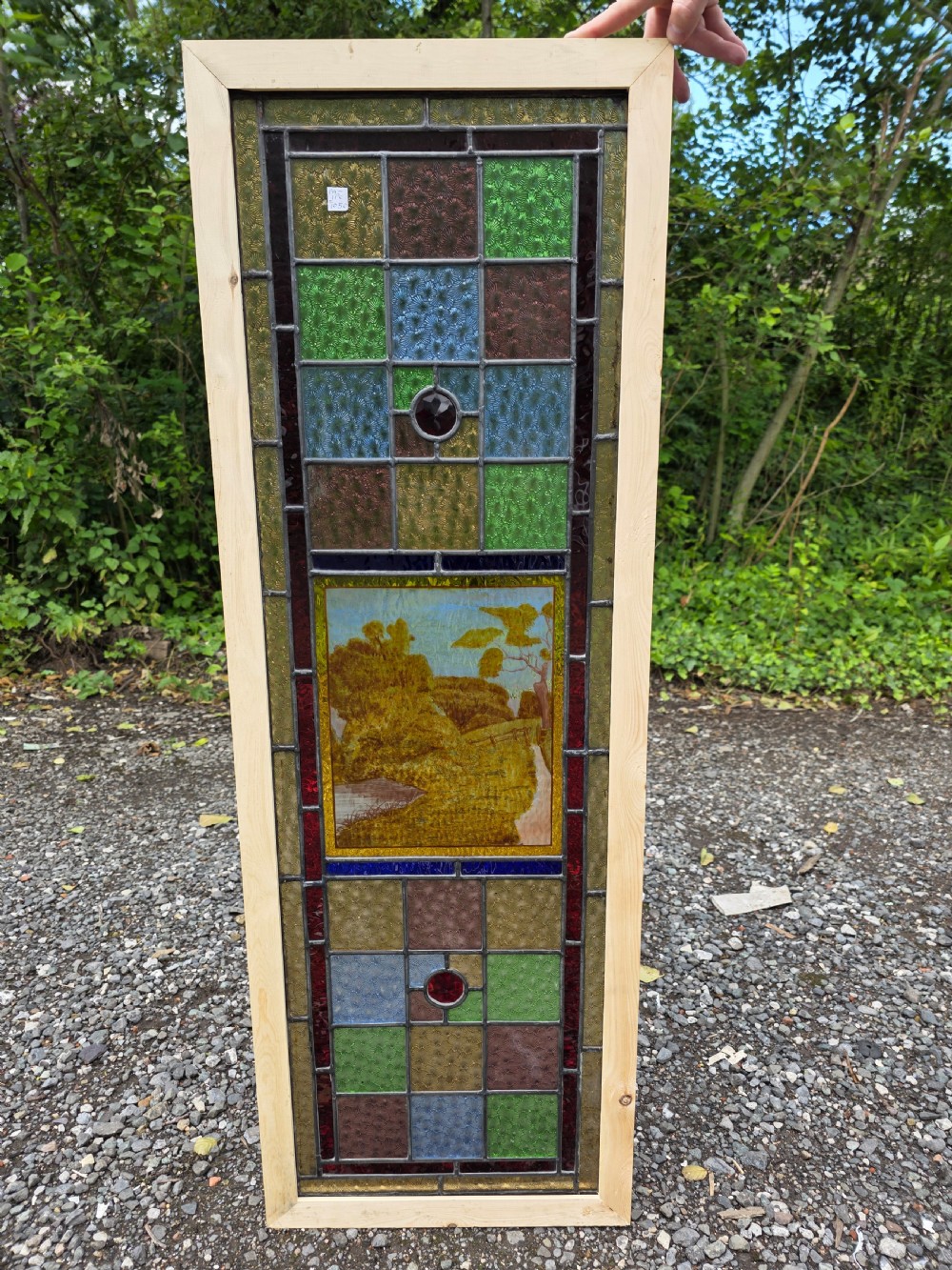 A narrow rectangular leaded glass window panel with geometric coloured panes, glass jewels, and a central hand-painted countryside scene. View IV
