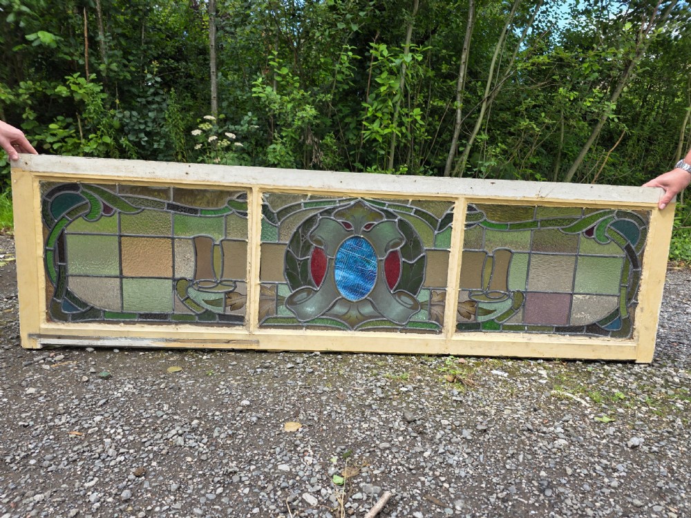 A wide rectangular antique leaded glass transom window featuring three sections of green, amber, blue, and red coloured glass with Art Nouveau scrollwork motifs set in a timber frame.