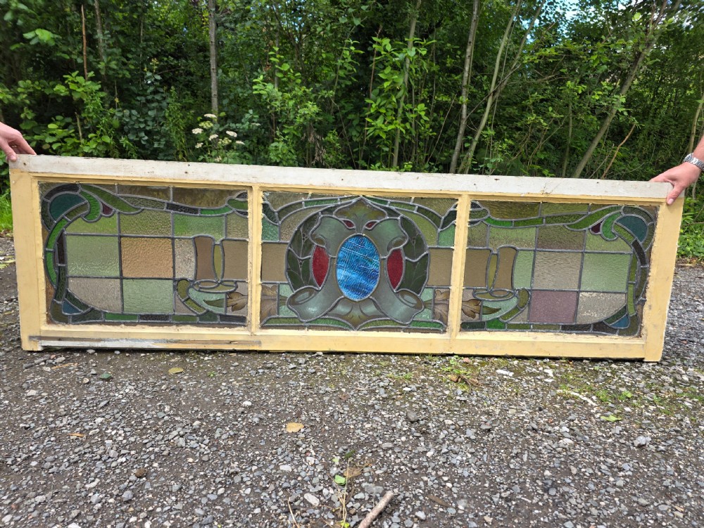 A wide rectangular antique leaded glass transom window featuring three sections of green, amber, blue, and red coloured glass with Art Nouveau scrollwork motifs set in a timber frame. View II