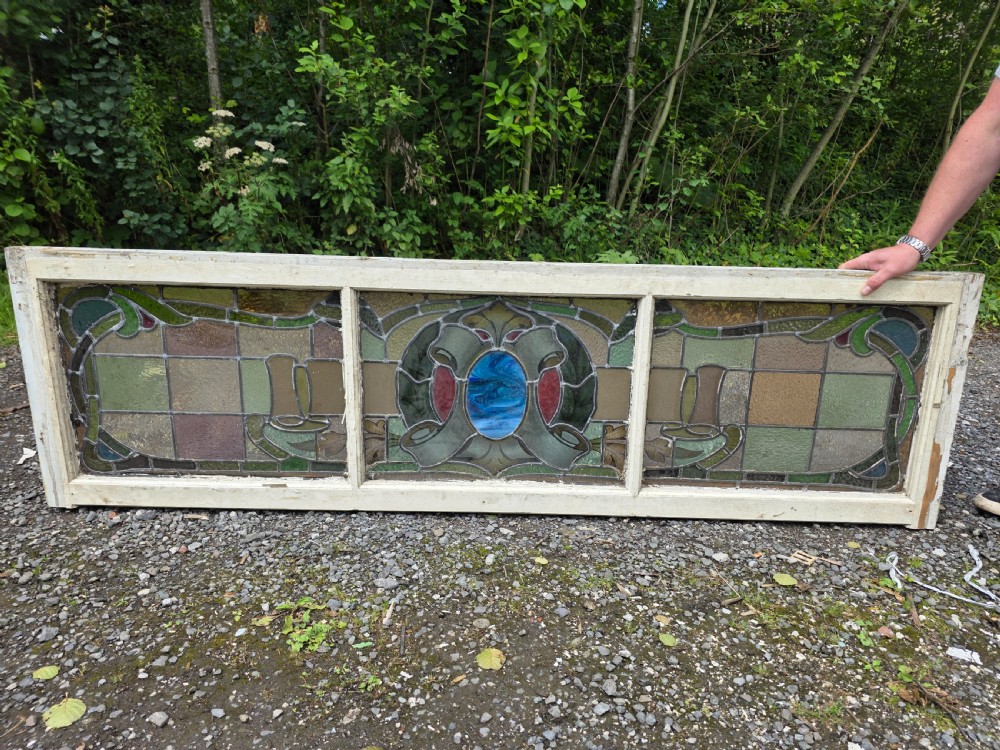 A wide rectangular antique leaded glass transom window featuring three sections of green, amber, blue, and red coloured glass with Art Nouveau scrollwork motifs set in a timber frame. View III
