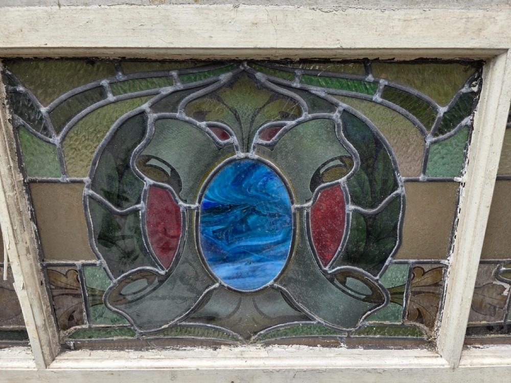 A wide rectangular antique leaded glass transom window featuring three sections of green, amber, blue, and red coloured glass with Art Nouveau scrollwork motifs set in a timber frame. View IV