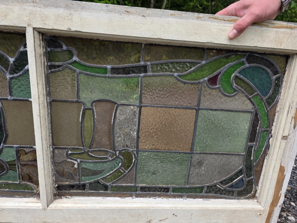 A wide rectangular antique leaded glass transom window featuring three sections of green, amber, blue, and red coloured glass with Art Nouveau scrollwork motifs set in a timber frame. View VI