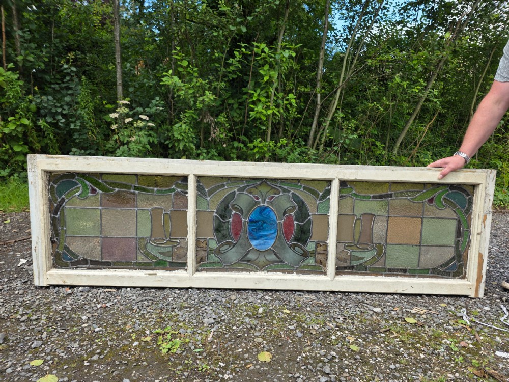 A wide rectangular antique leaded glass transom window featuring three sections of green, amber, blue, and red coloured glass with Art Nouveau scrollwork motifs set in a timber frame. View VII