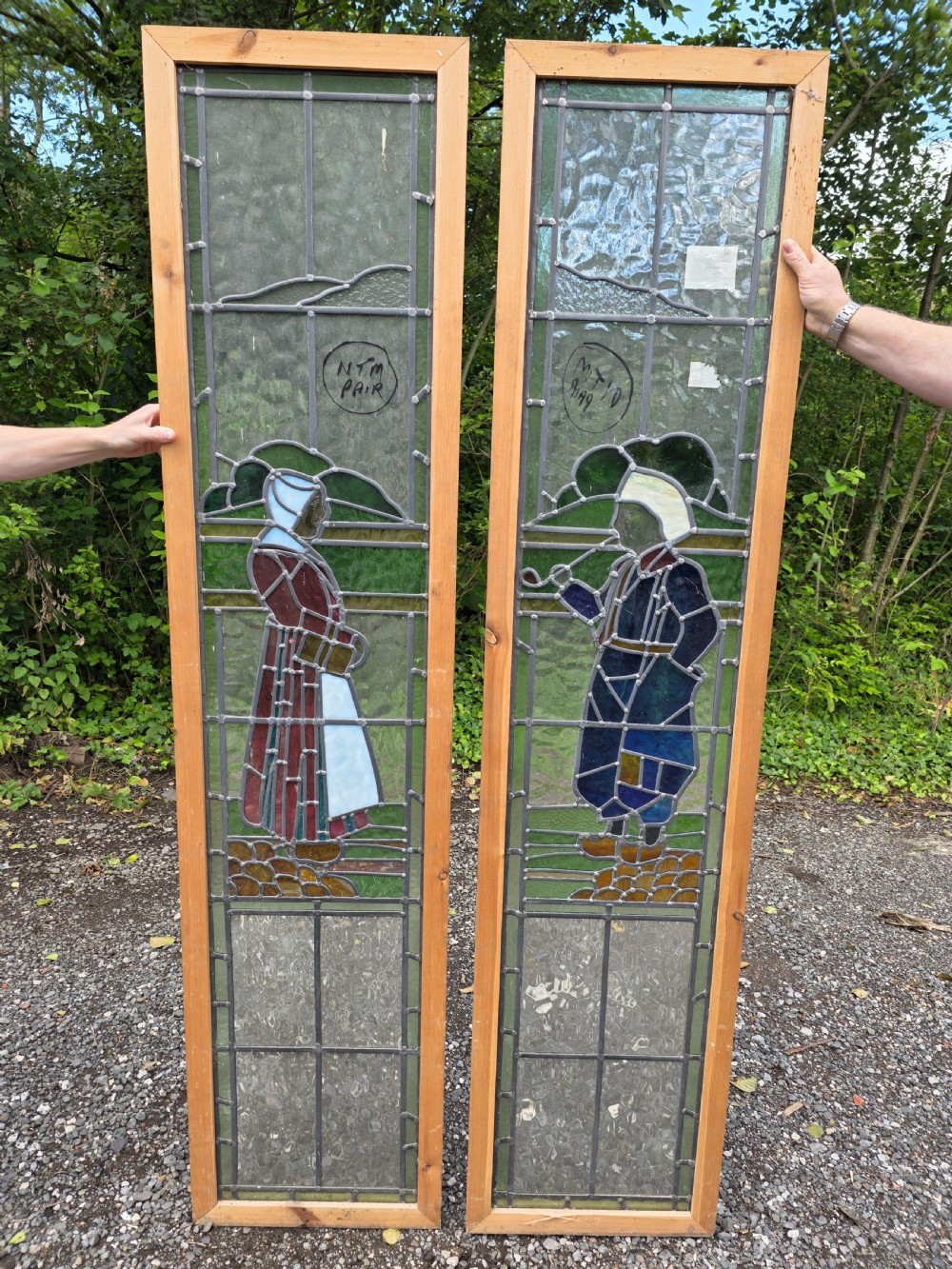 A pair of tall narrow leaded glass window panels set in light wood frames depicting a Dutch girl and boy in traditional dress against textured glass backdrop.