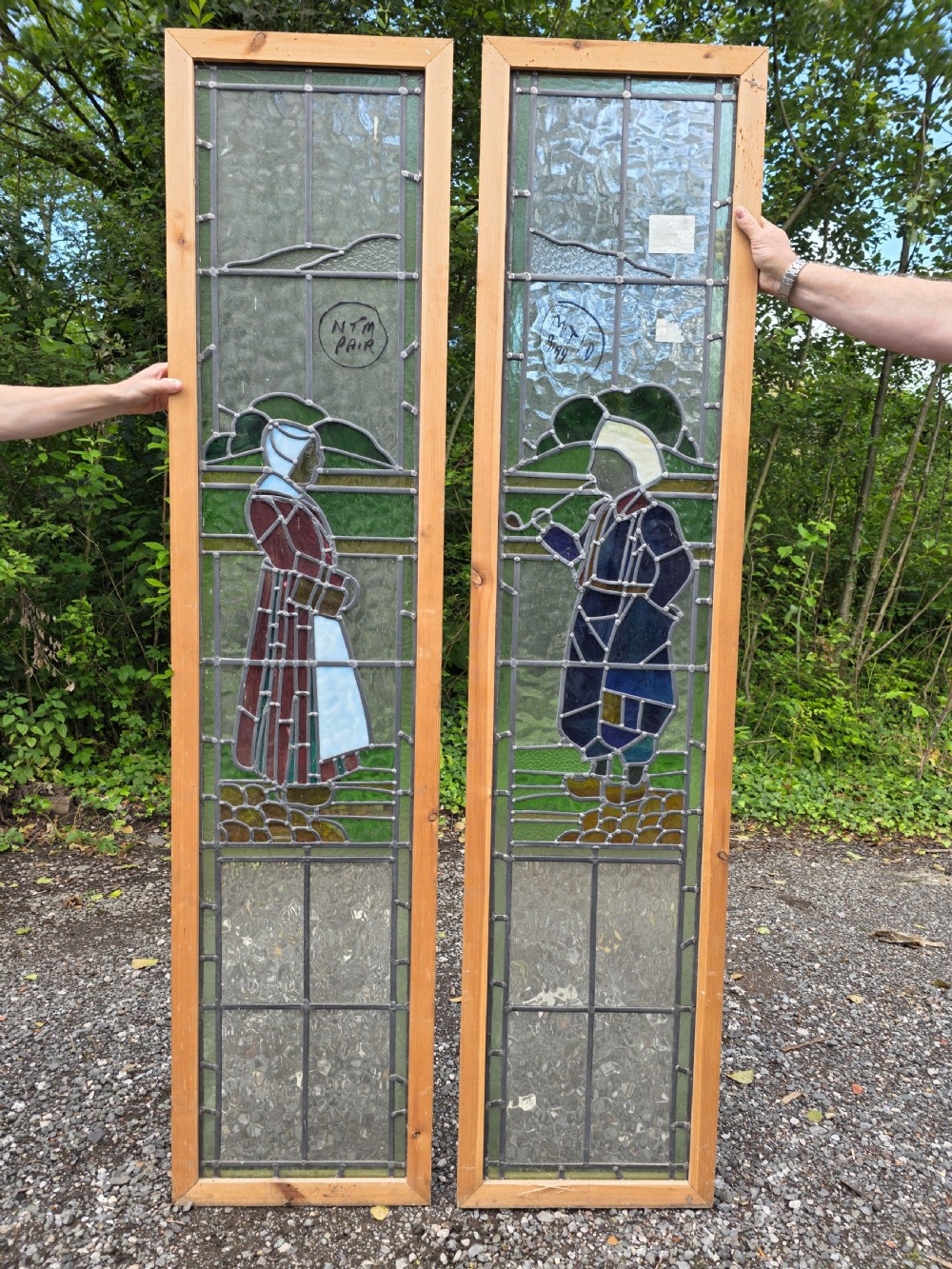 A pair of tall narrow leaded glass window panels set in light wood frames depicting a Dutch girl and boy in traditional dress against textured glass backdrop. View II