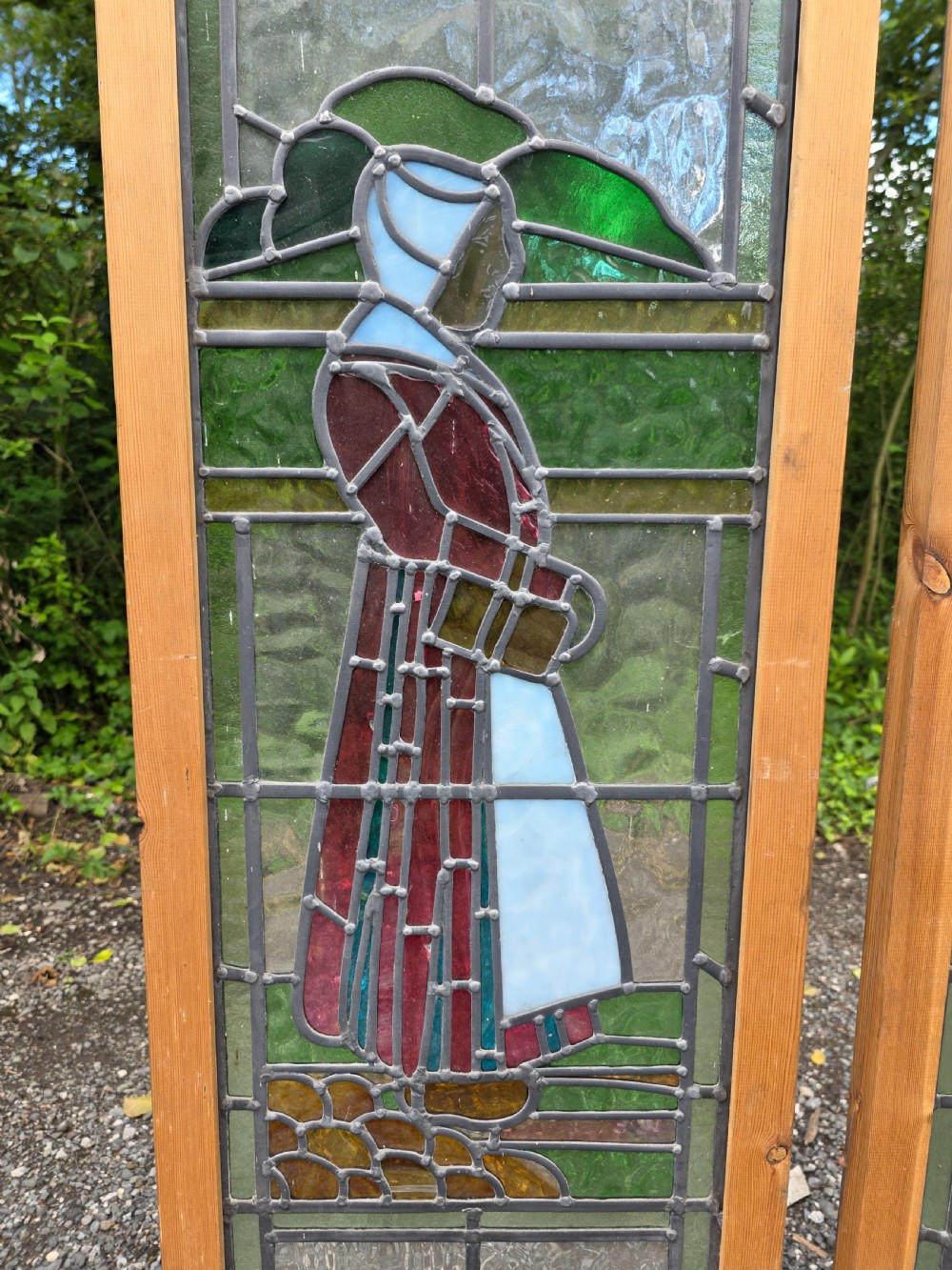 A pair of tall narrow leaded glass window panels set in light wood frames depicting a Dutch girl and boy in traditional dress against textured glass backdrop. View III