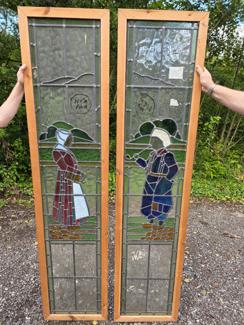 A pair of tall narrow leaded glass window panels set in light wood frames depicting a Dutch girl and boy in traditional dress against textured glass backdrop. View IV