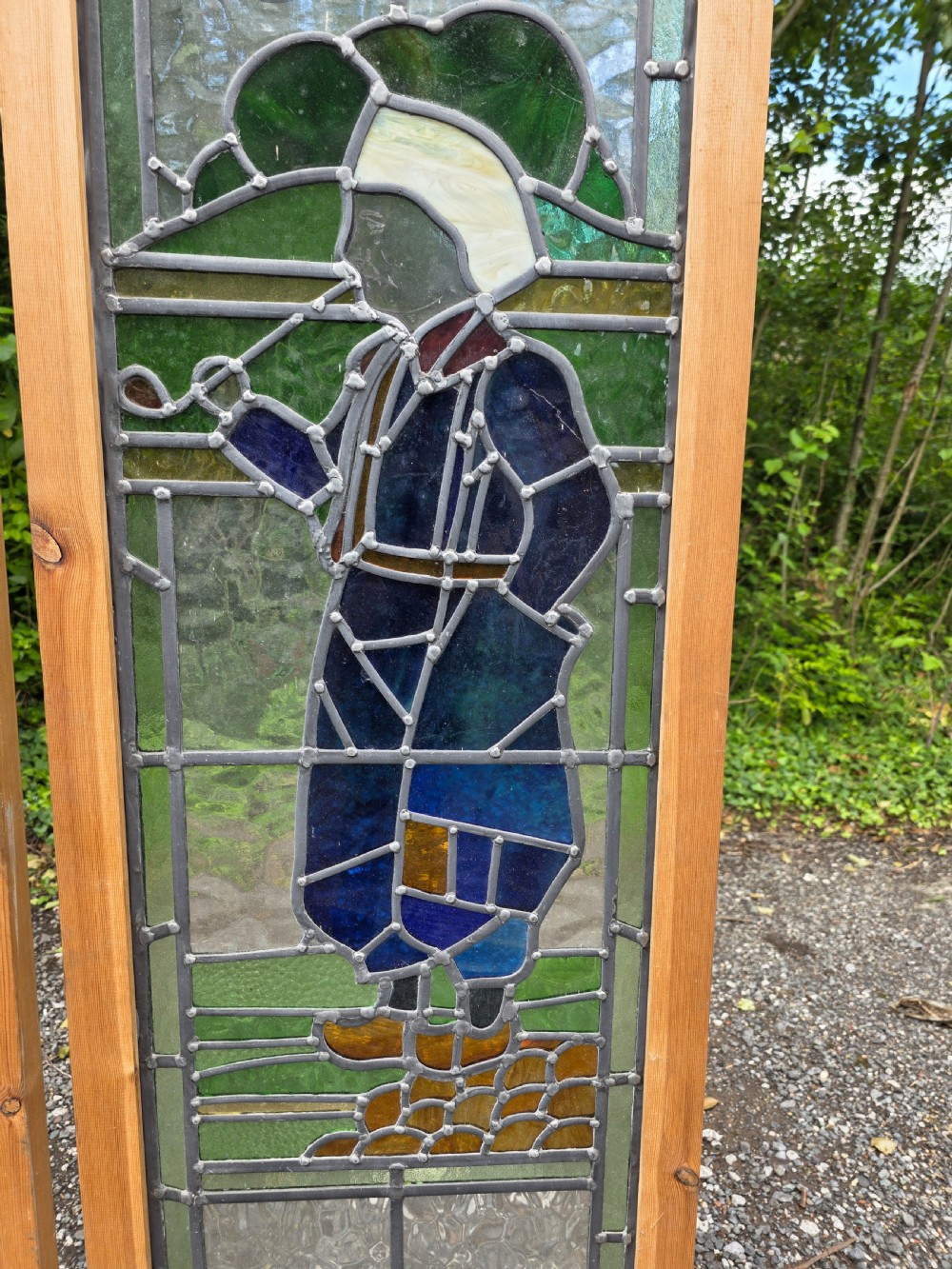 A pair of tall narrow leaded glass window panels set in light wood frames depicting a Dutch girl and boy in traditional dress against textured glass backdrop. View V