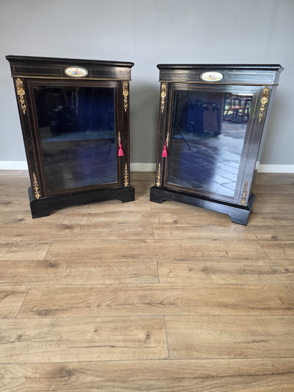 A pair of Victorian ebonised wooden pier cabinets featuring single glazed doors, gilt metal ormolu corner mounts, and oval porcelain plaques.