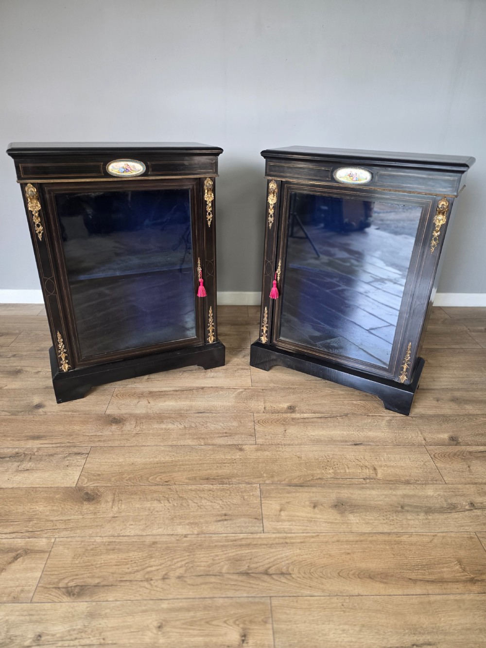 A pair of Victorian ebonised wooden pier cabinets featuring single glazed doors, gilt metal ormolu corner mounts, and oval porcelain plaques. View II