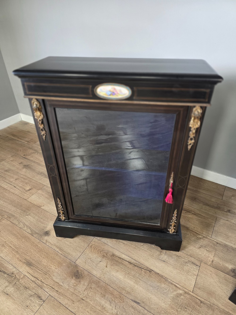 A pair of Victorian ebonised wooden pier cabinets featuring single glazed doors, gilt metal ormolu corner mounts, and oval porcelain plaques. View V