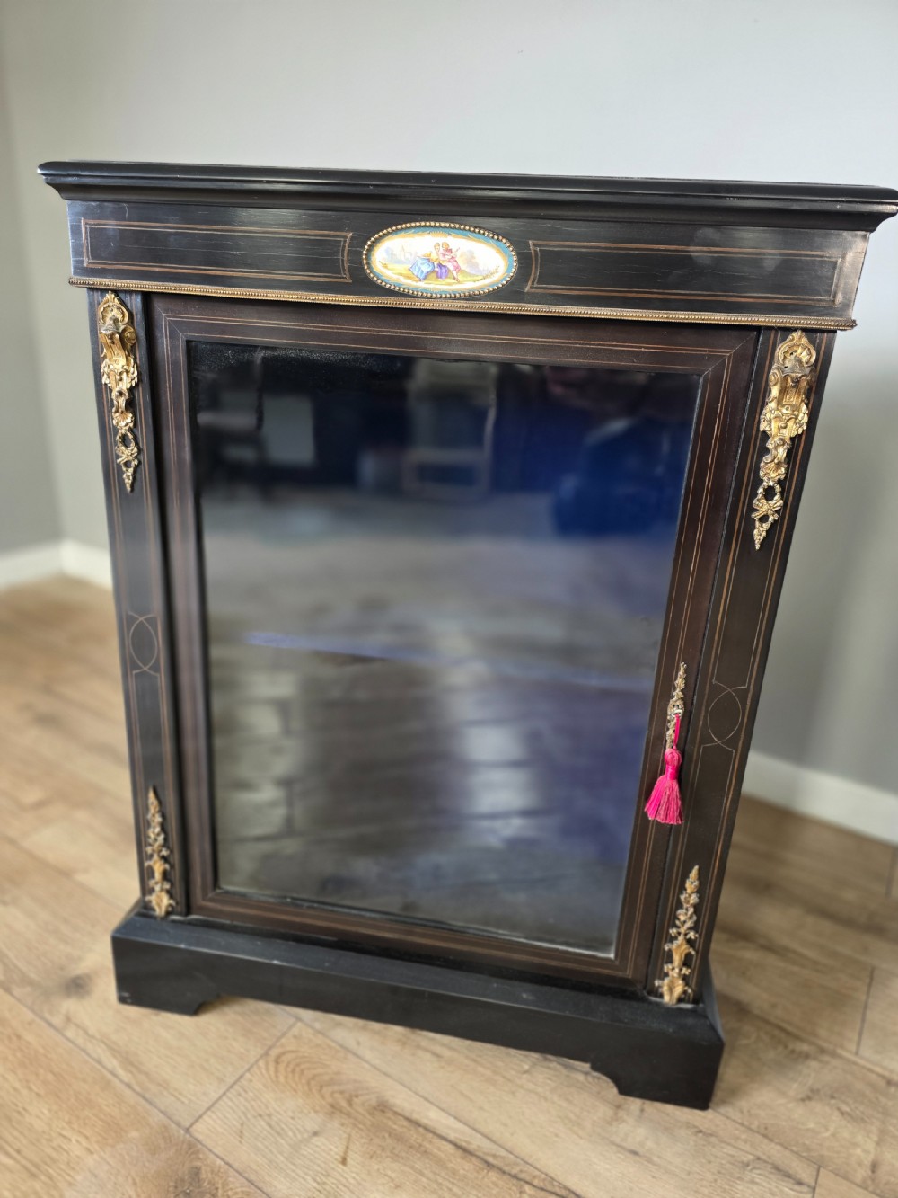 A pair of Victorian ebonised wooden pier cabinets featuring single glazed doors, gilt metal ormolu corner mounts, and oval porcelain plaques. View VI