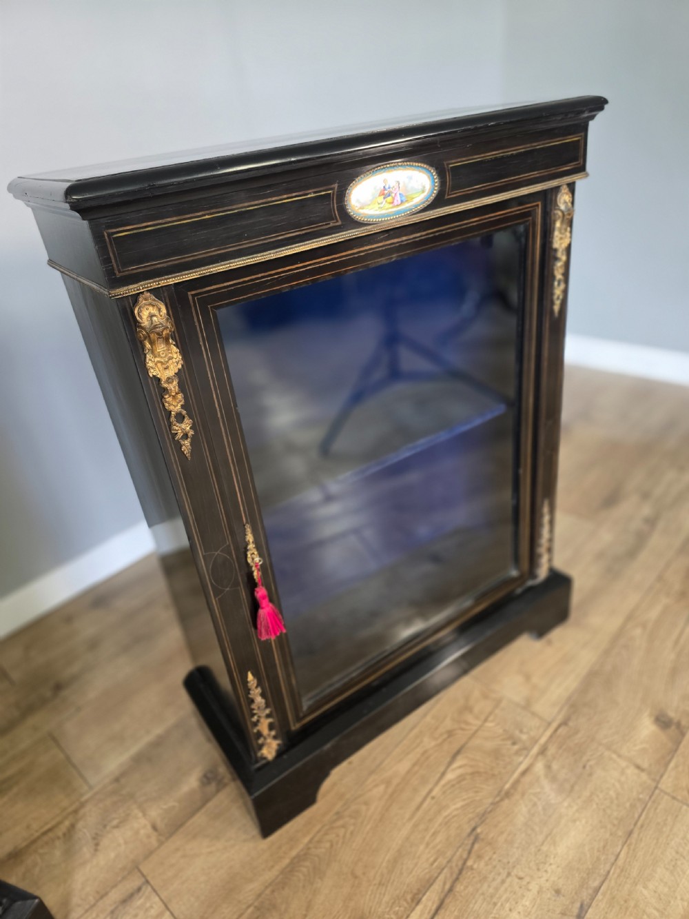 A pair of Victorian ebonised wooden pier cabinets featuring single glazed doors, gilt metal ormolu corner mounts, and oval porcelain plaques. View IX