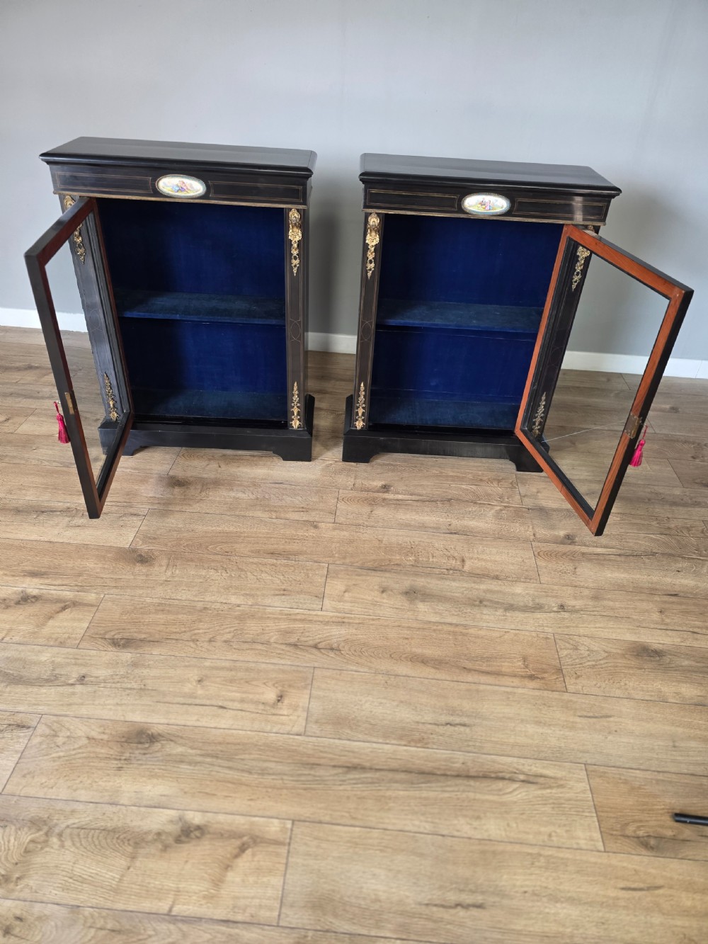A pair of Victorian ebonised wooden pier cabinets featuring single glazed doors, gilt metal ormolu corner mounts, and oval porcelain plaques. View X