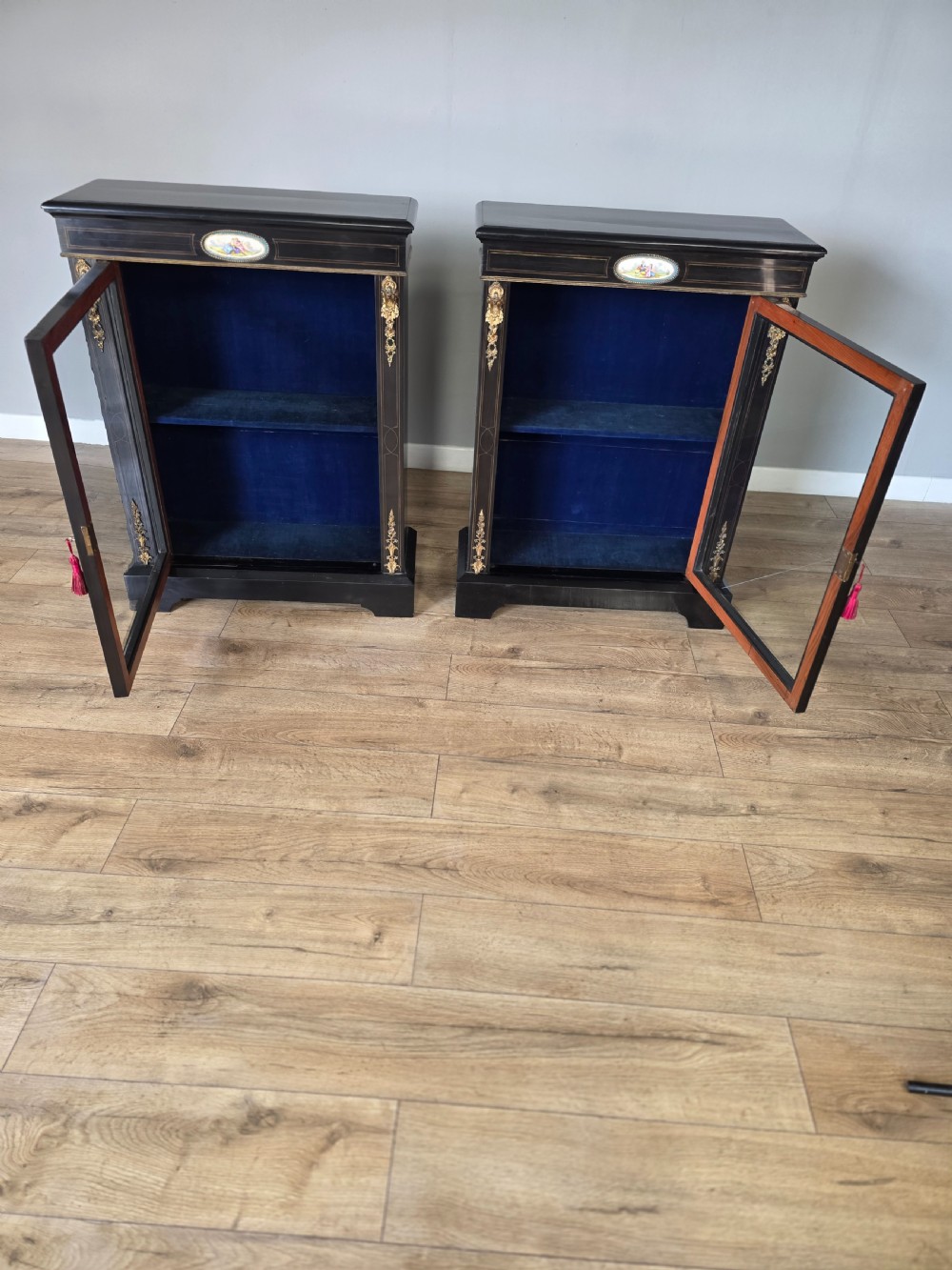 A pair of Victorian ebonised wooden pier cabinets featuring single glazed doors, gilt metal ormolu corner mounts, and oval porcelain plaques. View XI