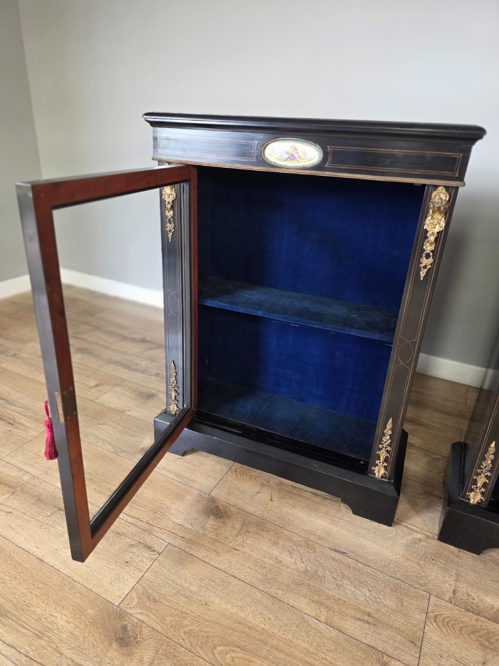 A pair of Victorian ebonised wooden pier cabinets featuring single glazed doors, gilt metal ormolu corner mounts, and oval porcelain plaques. View XII