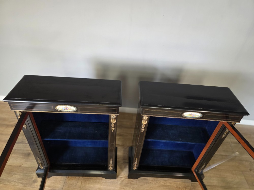 A pair of Victorian ebonised wooden pier cabinets featuring single glazed doors, gilt metal ormolu corner mounts, and oval porcelain plaques. View XV