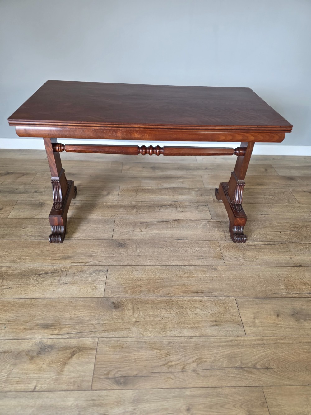 An early Victorian mahogany side table featuring a rectangular top, a central turned stretcher, and carved trestle feet resting on a light wood floor.