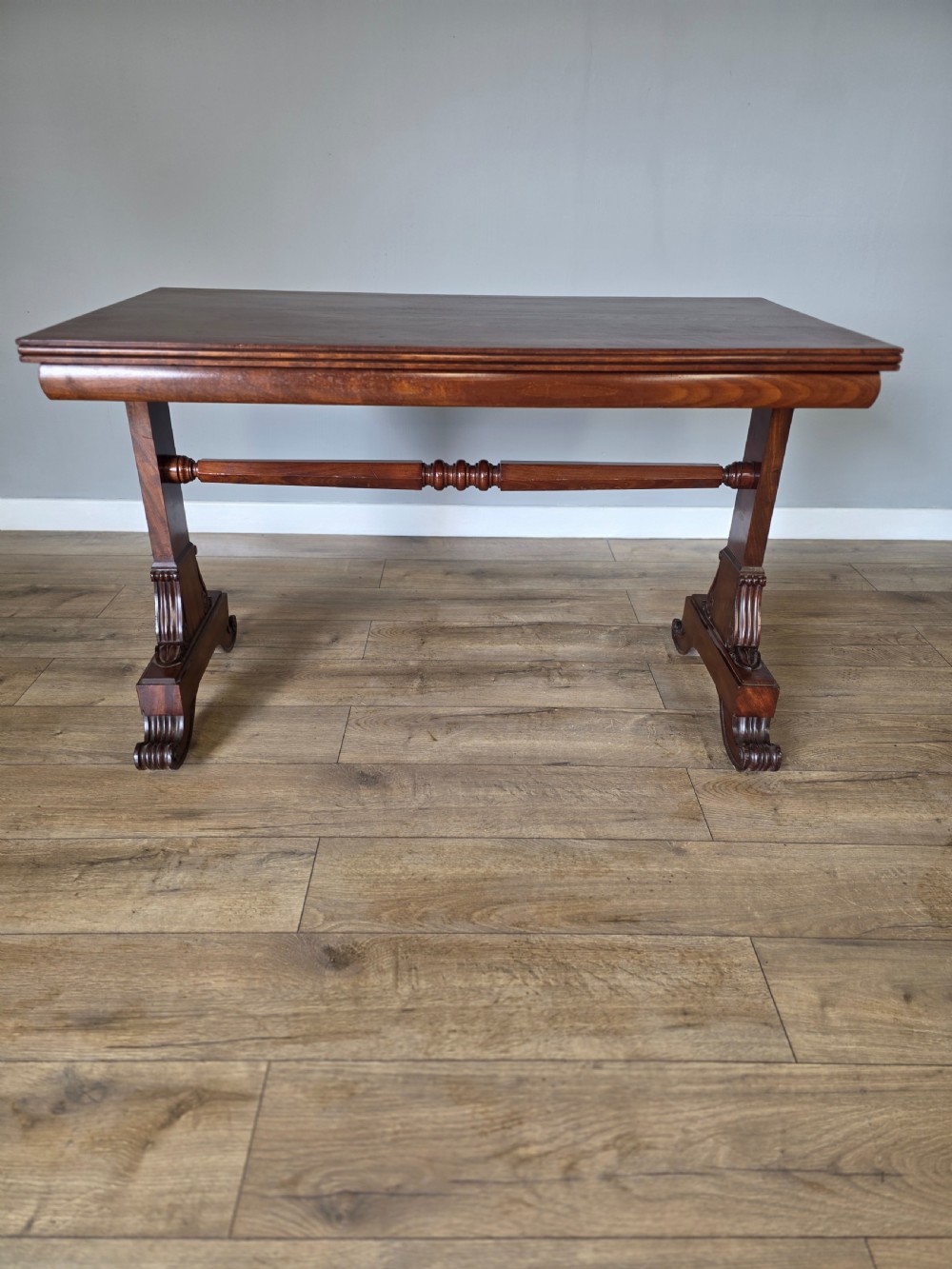 An early Victorian mahogany side table featuring a rectangular top, a central turned stretcher, and carved trestle feet resting on a light wood floor. View II