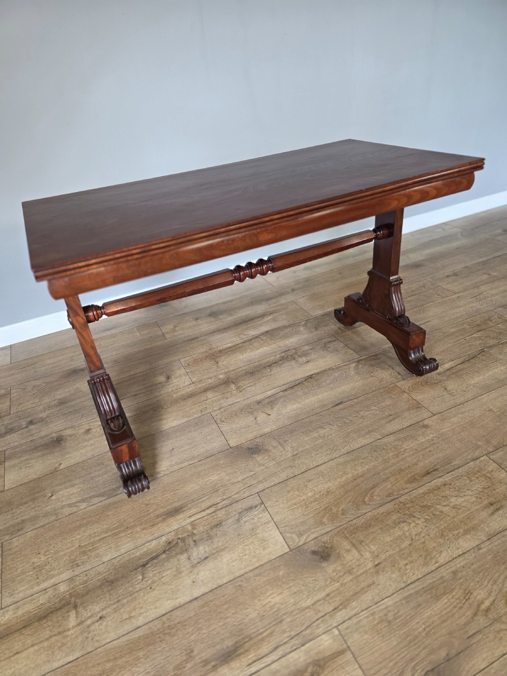 An early Victorian mahogany side table featuring a rectangular top, a central turned stretcher, and carved trestle feet resting on a light wood floor. View IV