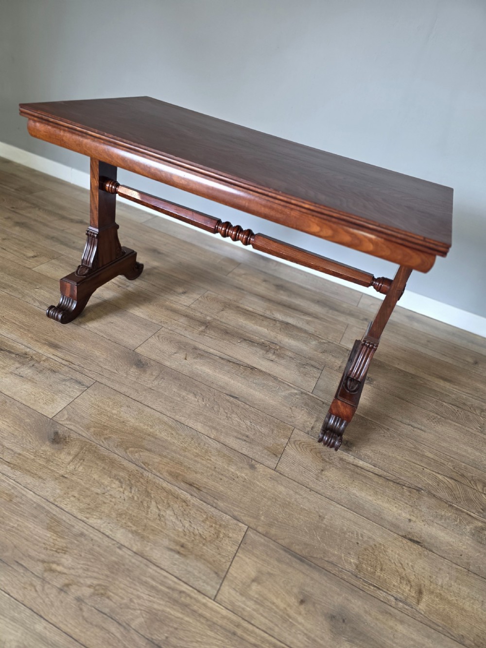 An early Victorian mahogany side table featuring a rectangular top, a central turned stretcher, and carved trestle feet resting on a light wood floor. View V