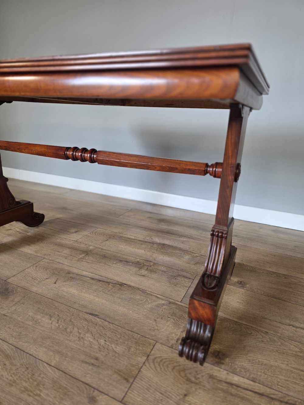 An early Victorian mahogany side table featuring a rectangular top, a central turned stretcher, and carved trestle feet resting on a light wood floor. View VI