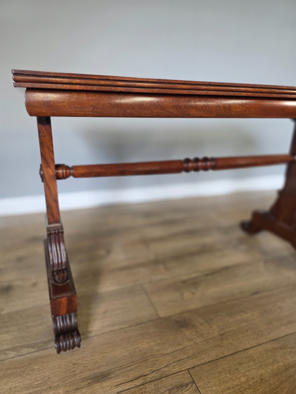 An early Victorian mahogany side table featuring a rectangular top, a central turned stretcher, and carved trestle feet resting on a light wood floor. View VII