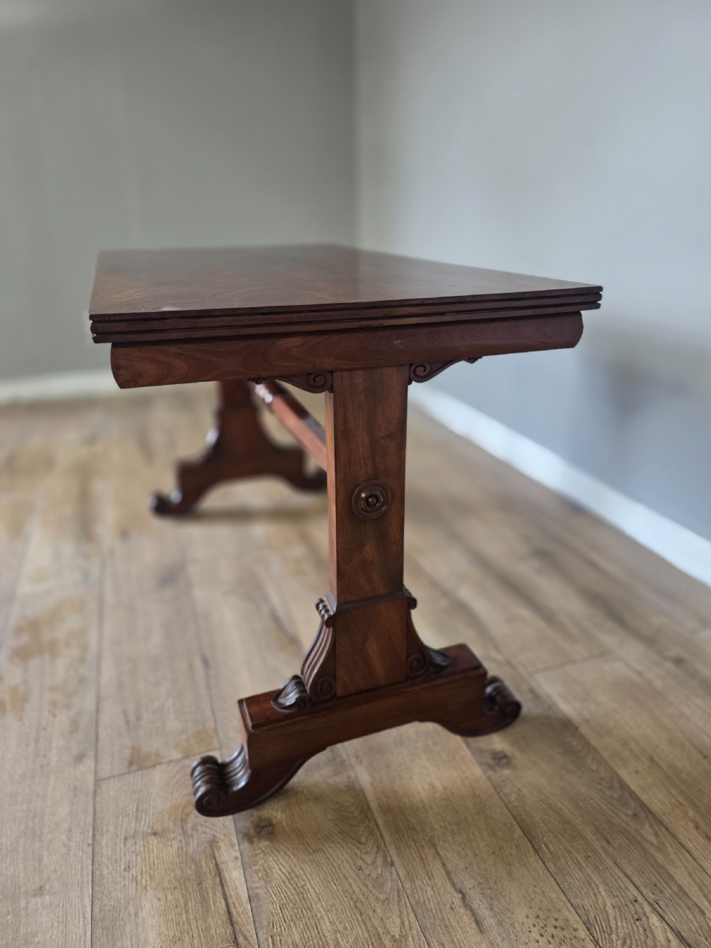 An early Victorian mahogany side table featuring a rectangular top, a central turned stretcher, and carved trestle feet resting on a light wood floor. View VIII