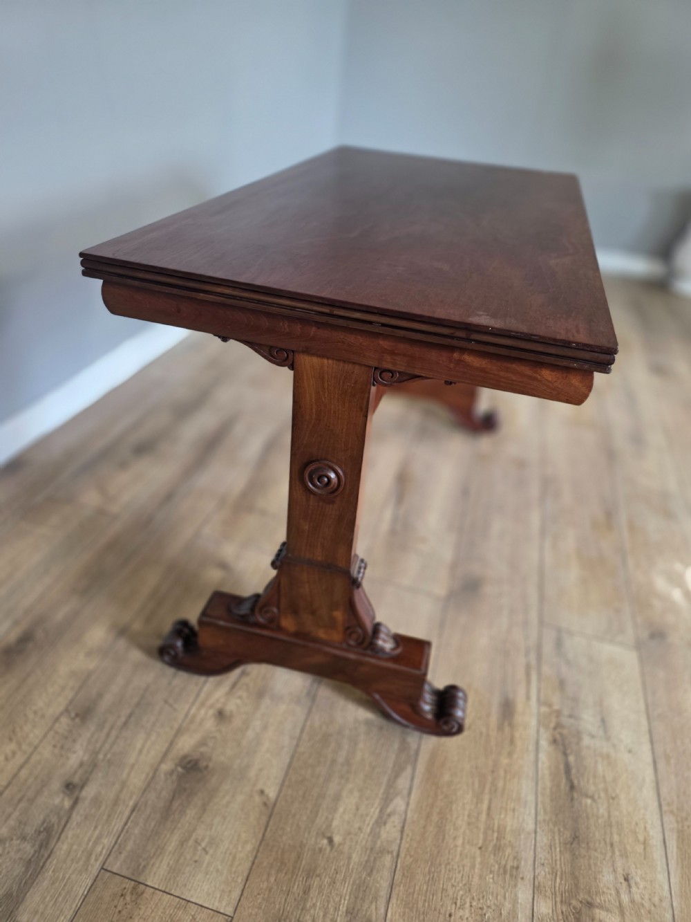 An early Victorian mahogany side table featuring a rectangular top, a central turned stretcher, and carved trestle feet resting on a light wood floor. View IX