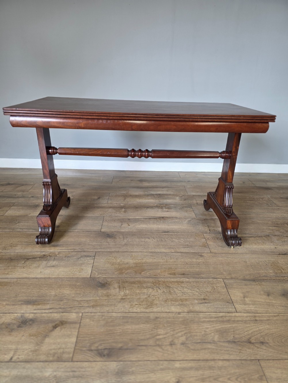 An early Victorian mahogany side table featuring a rectangular top, a central turned stretcher, and carved trestle feet resting on a light wood floor. View XII