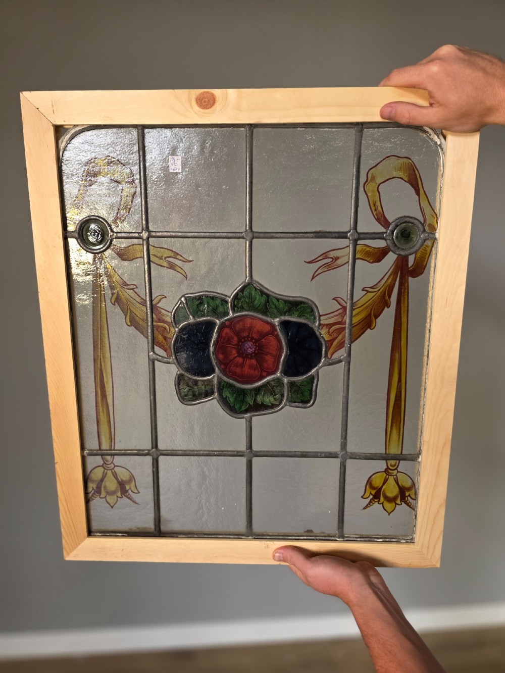An antique rectangular leaded glass window panel featuring hand-painted red flowers, green leaves, yellow ribbon swags, and small glass bullseyes set within a wooden frame.