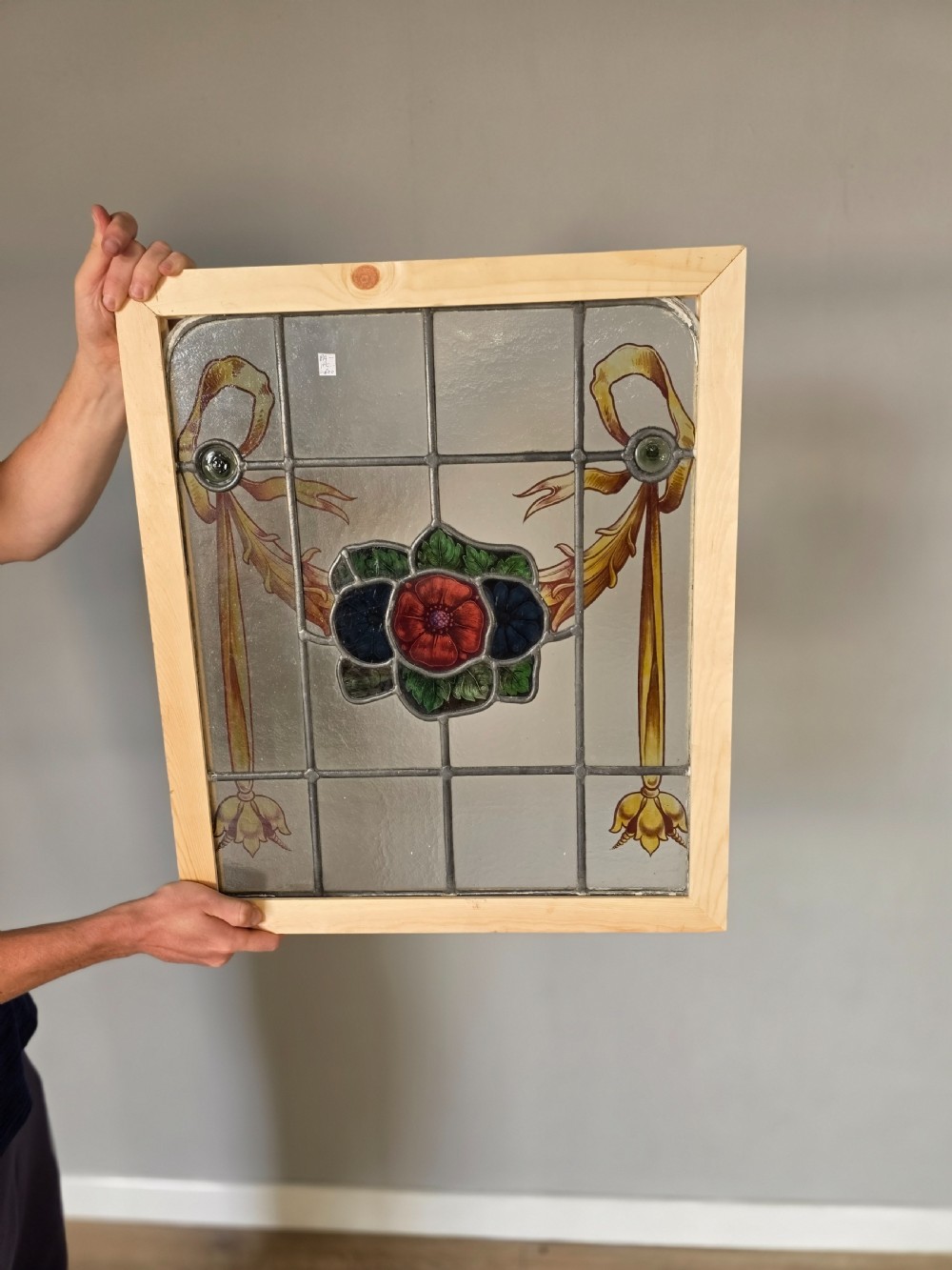 An antique rectangular leaded glass window panel featuring hand-painted red flowers, green leaves, yellow ribbon swags, and small glass bullseyes set within a wooden frame. View II