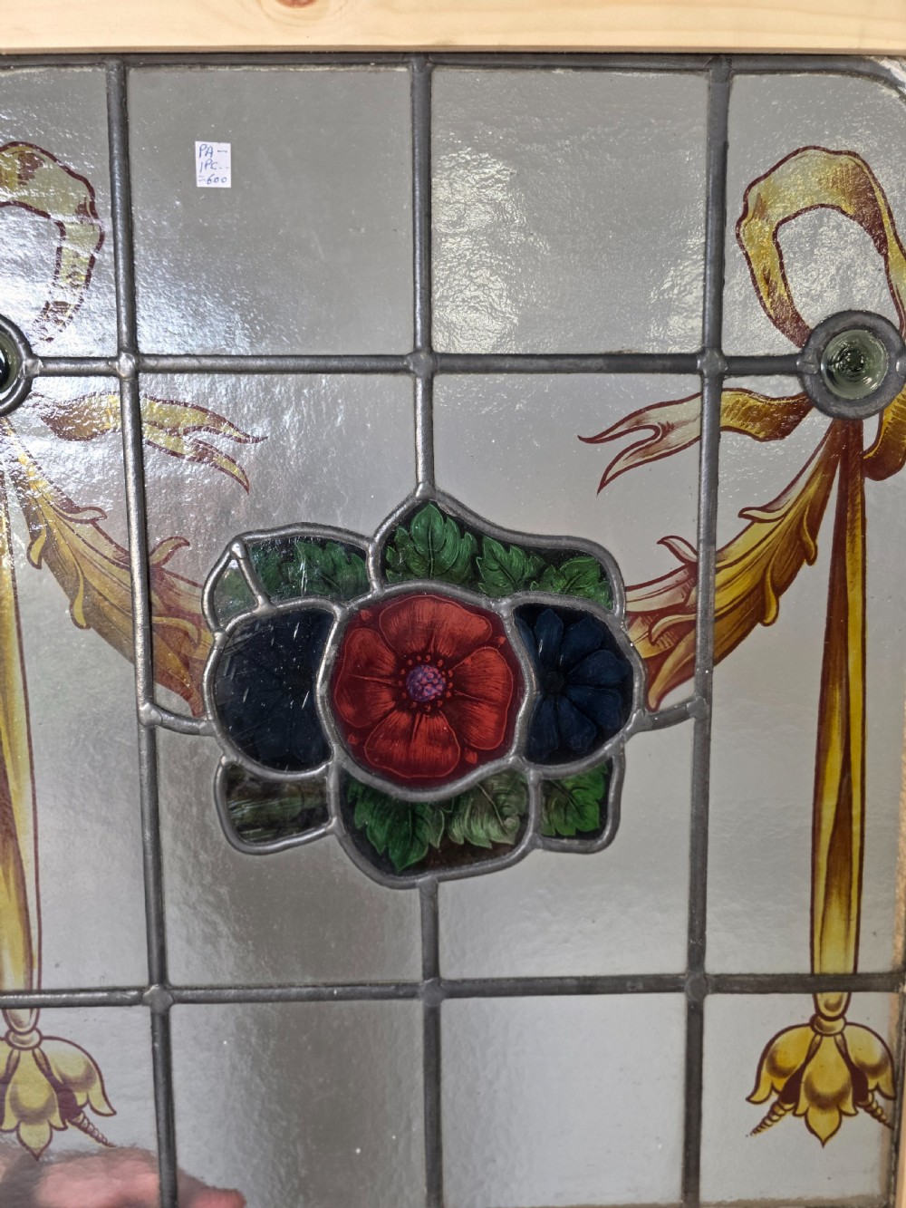 An antique rectangular leaded glass window panel featuring hand-painted red flowers, green leaves, yellow ribbon swags, and small glass bullseyes set within a wooden frame. View III