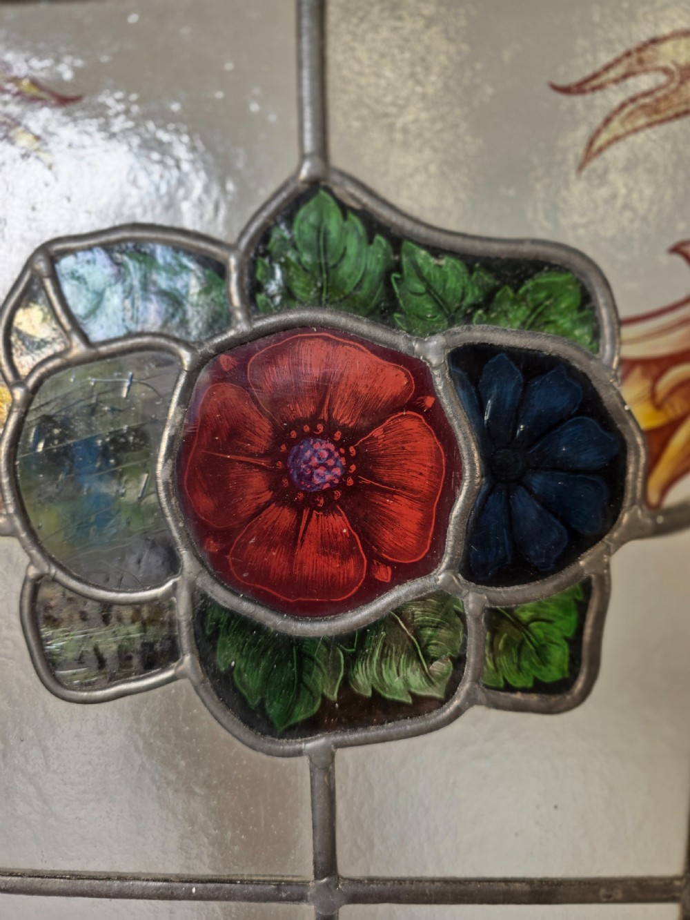 An antique rectangular leaded glass window panel featuring hand-painted red flowers, green leaves, yellow ribbon swags, and small glass bullseyes set within a wooden frame. View V