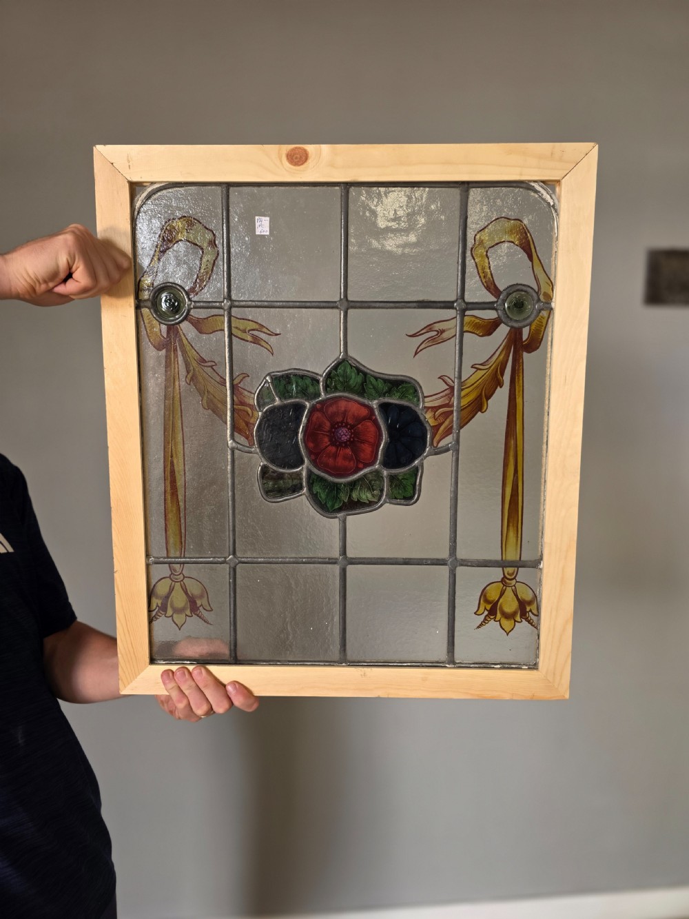 An antique rectangular leaded glass window panel featuring hand-painted red flowers, green leaves, yellow ribbon swags, and small glass bullseyes set within a wooden frame. View VI