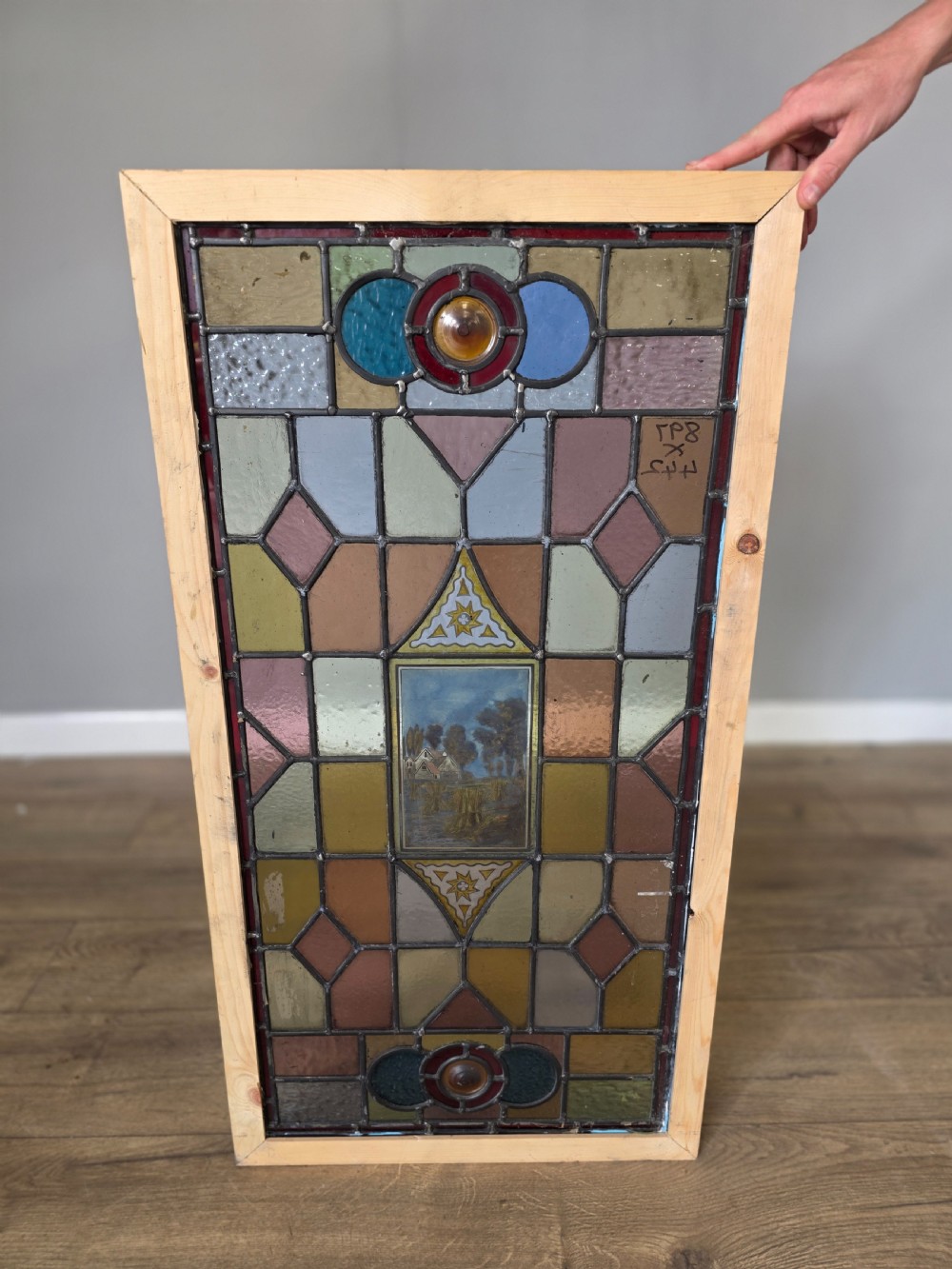 A rectangular leaded stained glass panel featuring geometric multicoloured glass sections and a central hand-painted country landscape.