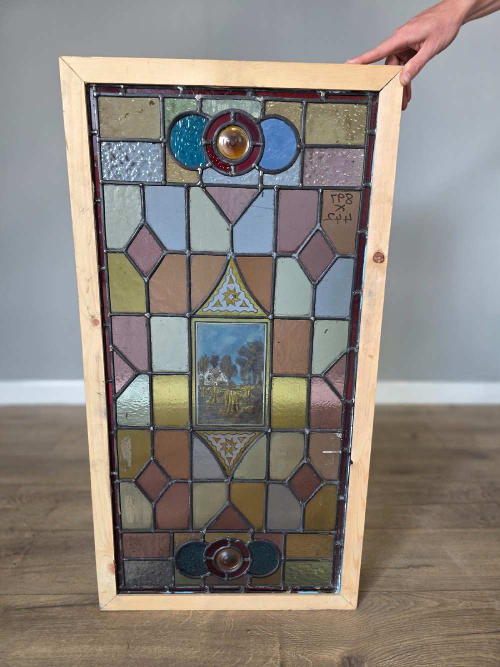 A rectangular leaded stained glass panel featuring geometric multicoloured glass sections and a central hand-painted country landscape. View V