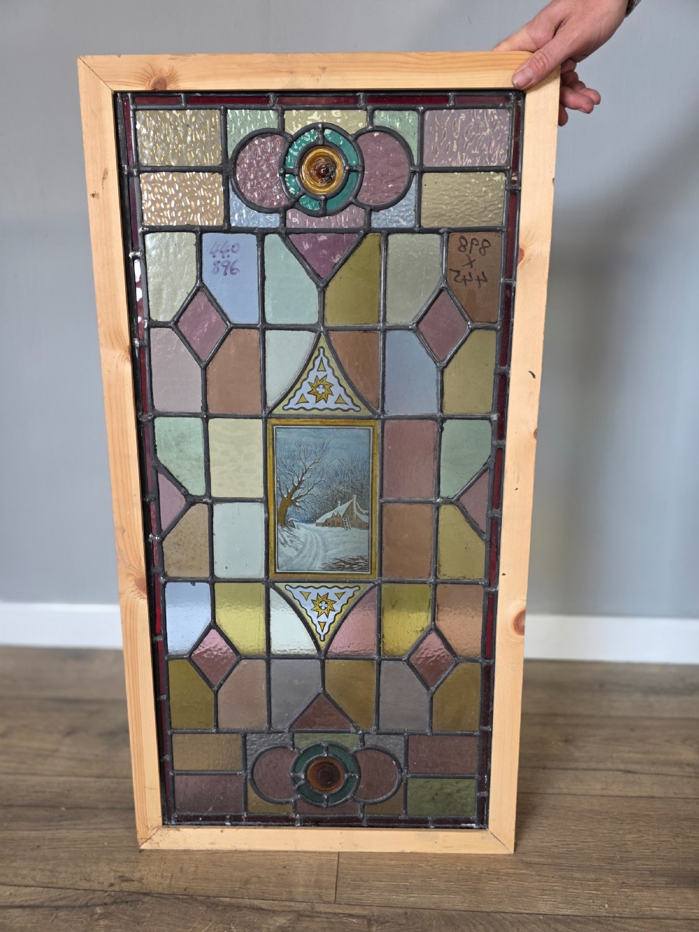 A rectangular Edwardian leaded stained glass window panel featuring geometric coloured glass, roundels, and a central hand-painted winter landscape scene set in a pine frame.