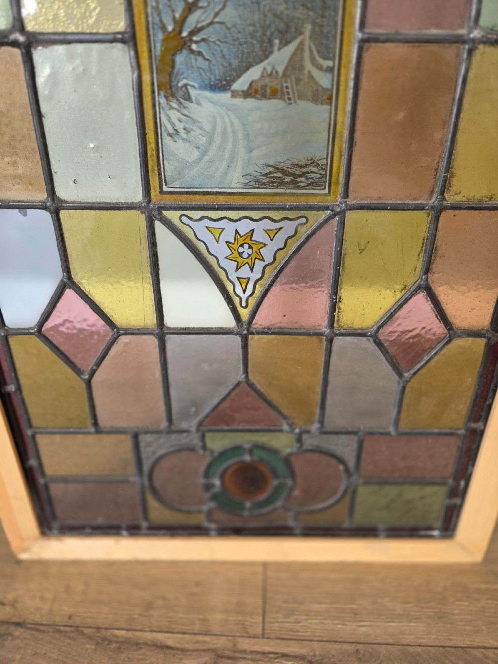 A rectangular Edwardian leaded stained glass window panel featuring geometric coloured glass, roundels, and a central hand-painted winter landscape scene set in a pine frame. View IV