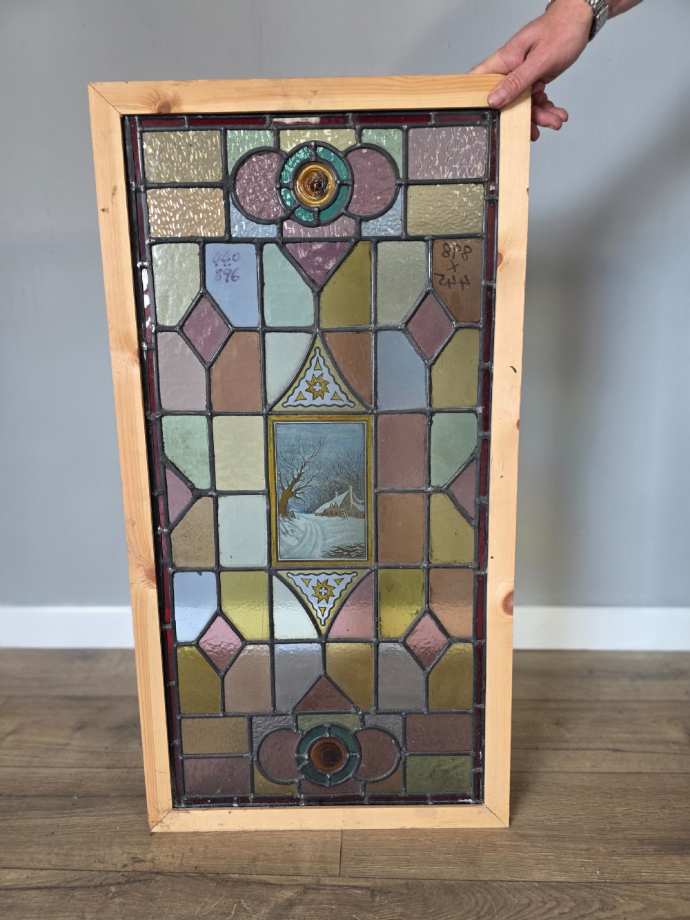 A rectangular Edwardian leaded stained glass window panel featuring geometric coloured glass, roundels, and a central hand-painted winter landscape scene set in a pine frame. View V