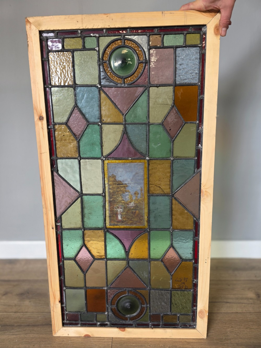 An antique rectangular leaded glass window panel featuring geometric coloured glass panes, bullseye roundels, and a central hand-painted countryside landscape vignette.