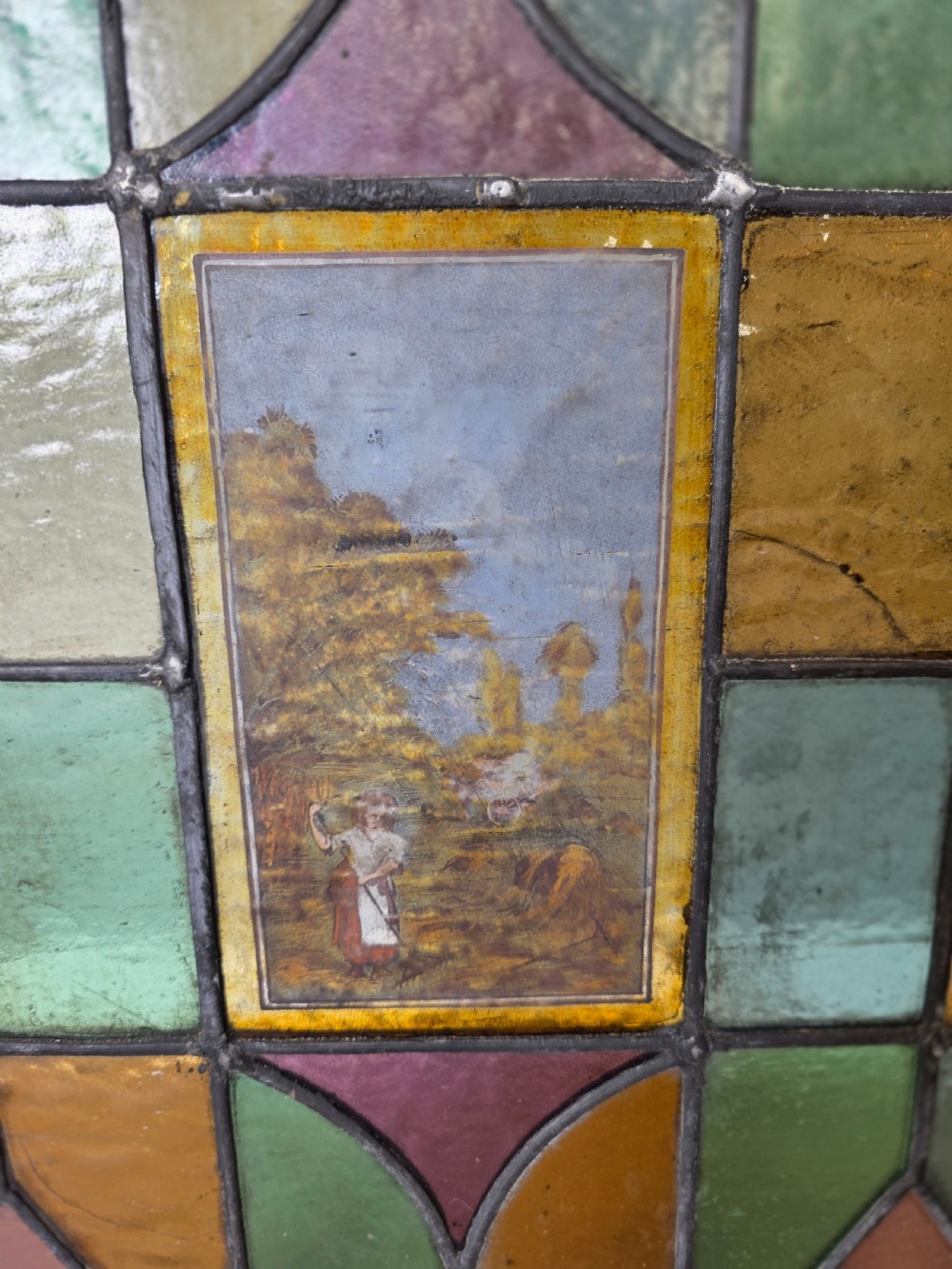 An antique rectangular leaded glass window panel featuring geometric coloured glass panes, bullseye roundels, and a central hand-painted countryside landscape vignette. View II